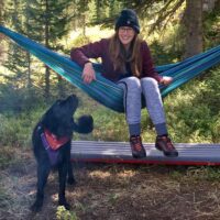 Aikens Ellen Aikens sits in a blue hammock in a green forest environment. A black dog looks back at her as if to say, "What are you doing?"