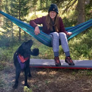 Ellen Aikens sits in a blue hammock in a green forest environment. A black dog looks back at her as if to say, "What are you doing?"