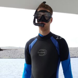 Mitch stands in a wetsuit, wearing goggles and a snorkel. Behind him is water and a bird flying by.