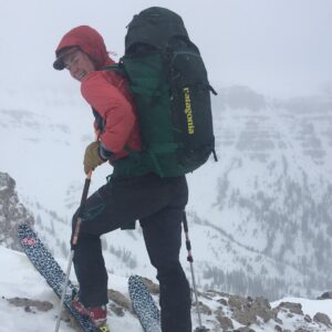 Sam Dwinnel stands with her back to the viewer, looking back and smiling. She appears to be at the edge of a snowy, rocky slope and is wearing skis and a large green backpack.
