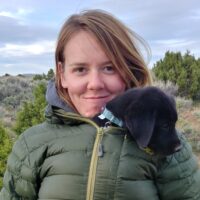 Katey Huggler stands in a sagebrush steppe environment, smiling at the camera. Her small puppy is zipped into her green jacket with her.