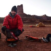 Alex May stirs a skillet full of food on a camp stove with tents and red rocks in the background.