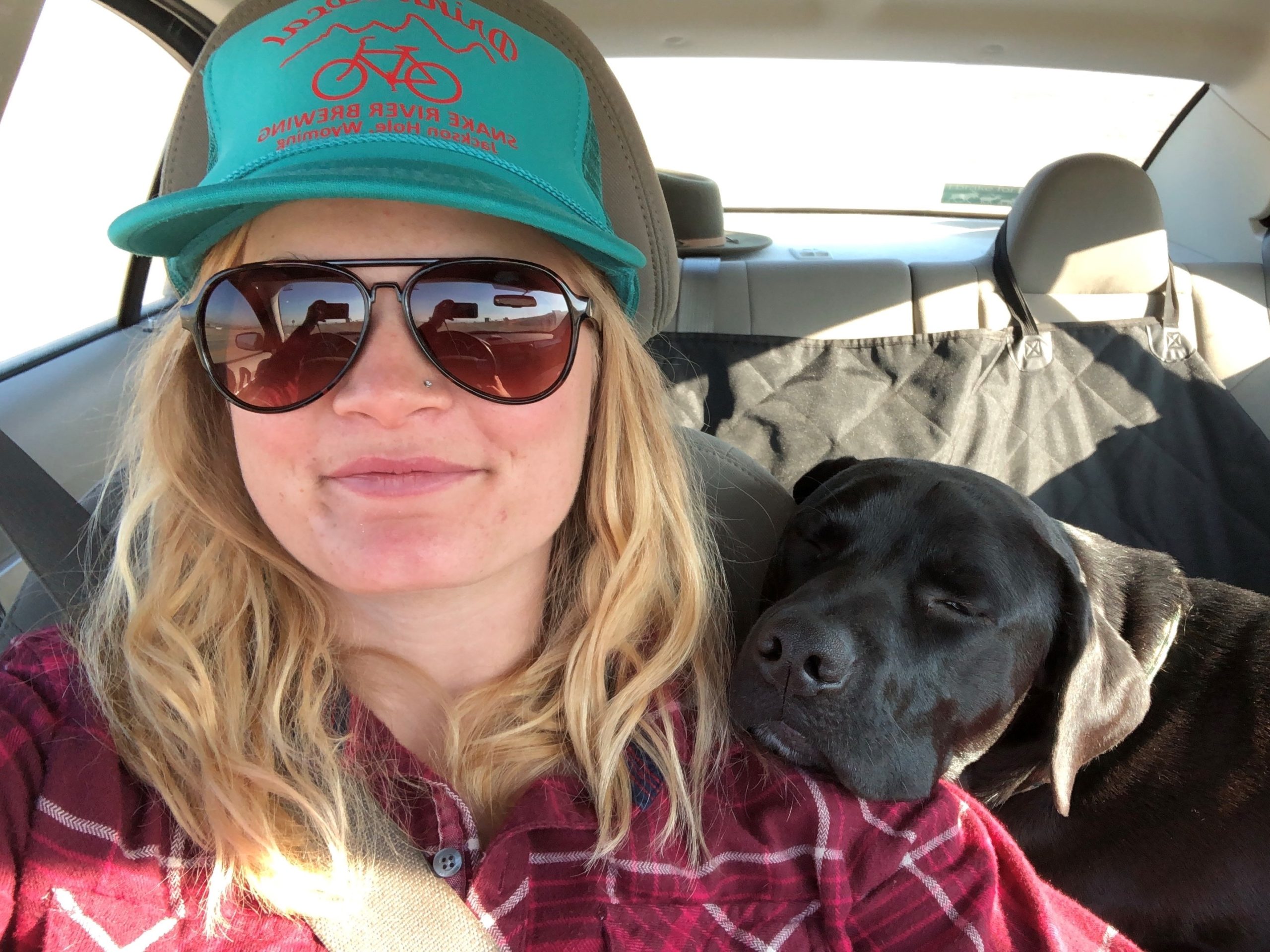Emily Reed smiles at the camera in a selfie from her car. Her pup leans their head on her shoulder.