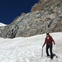 Smiley Rachel stands in a field of snow, using a pole to help balance her. Behind her is a very rocky outcrop, showing that she is in very high elevation, steep terrain.