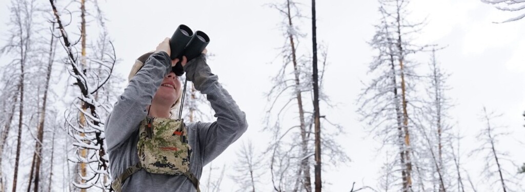 A scientist looks through binoculars at an unknown object. She is smiling. She is standing in a snowy forest, with burned trees covered in snow in the background.