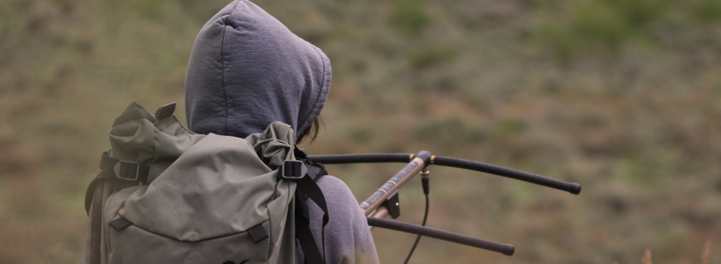 A scientist holds radio telemetry, searching for an animal in a sea of sagebrush. She wears a large backpack, and has her hood up.