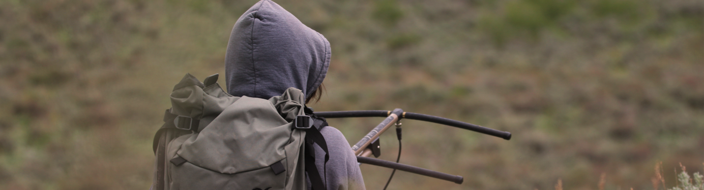 A scientist holds radio telemetry, searching for an animal in a sea of sagebrush. She wears a large backpack, and has her hood up.