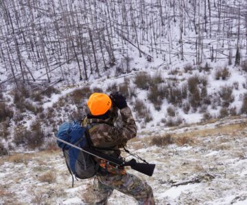 An orange-and-camouflage clad figure looks through binoculars in a snowy forest of dead trees.
