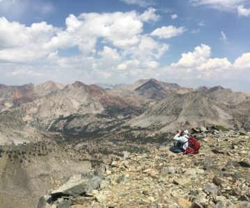 A person sits, wearing a backpack and looking through binoculars from the top of a rocky mountain: below, rocky slopes, forest, and a lake are shown.