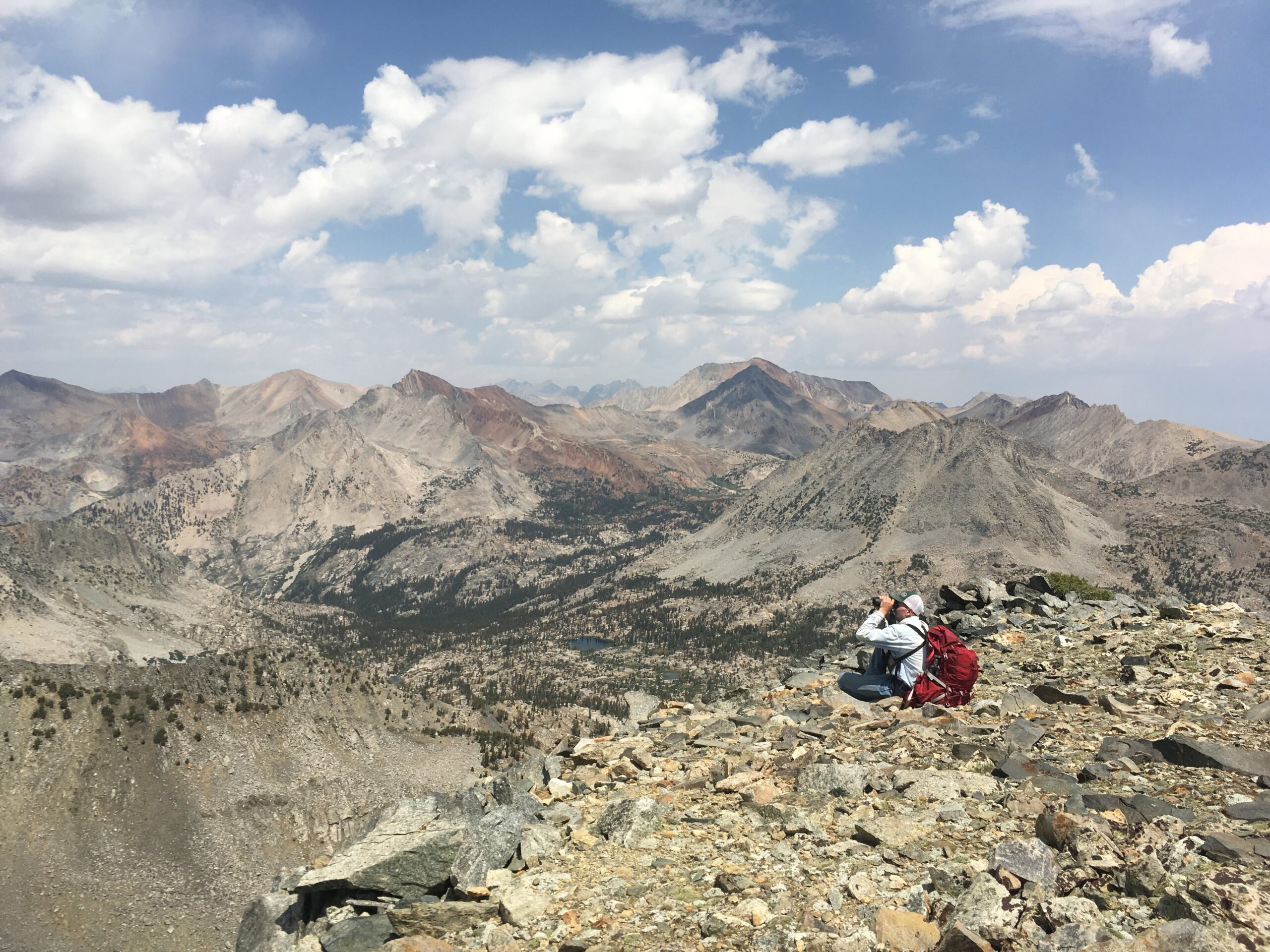 A person sits, wearing a backpack and looking through binoculars from the top of a rocky mountain: below, rocky slopes, forest, and a lake are shown.